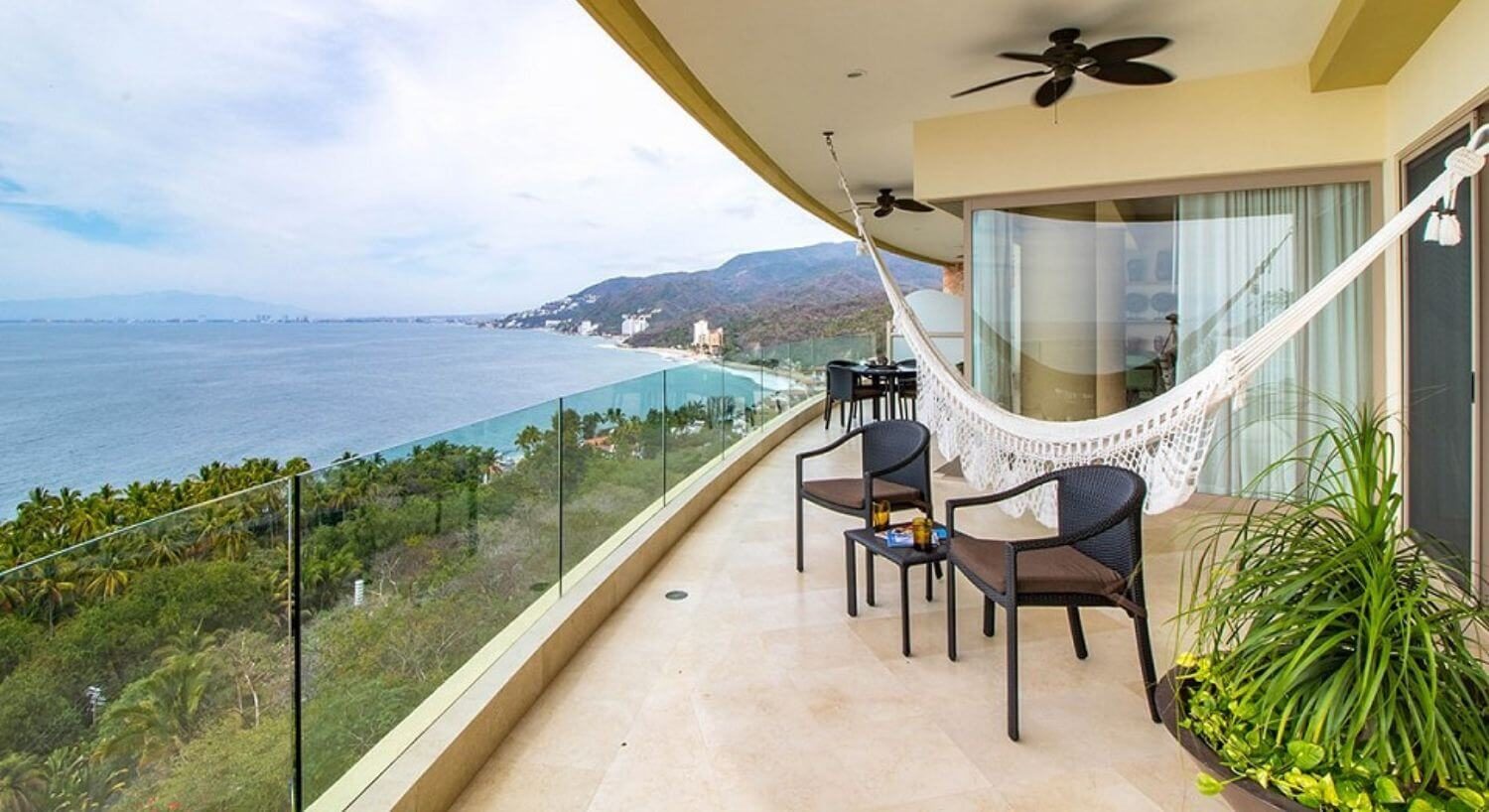 A private large balcony with patio furniture, a planter with green plants, a hammock, and dining table and chairs, overlooking the lush green mountainside, palm trees, ocean and nearby mountains.