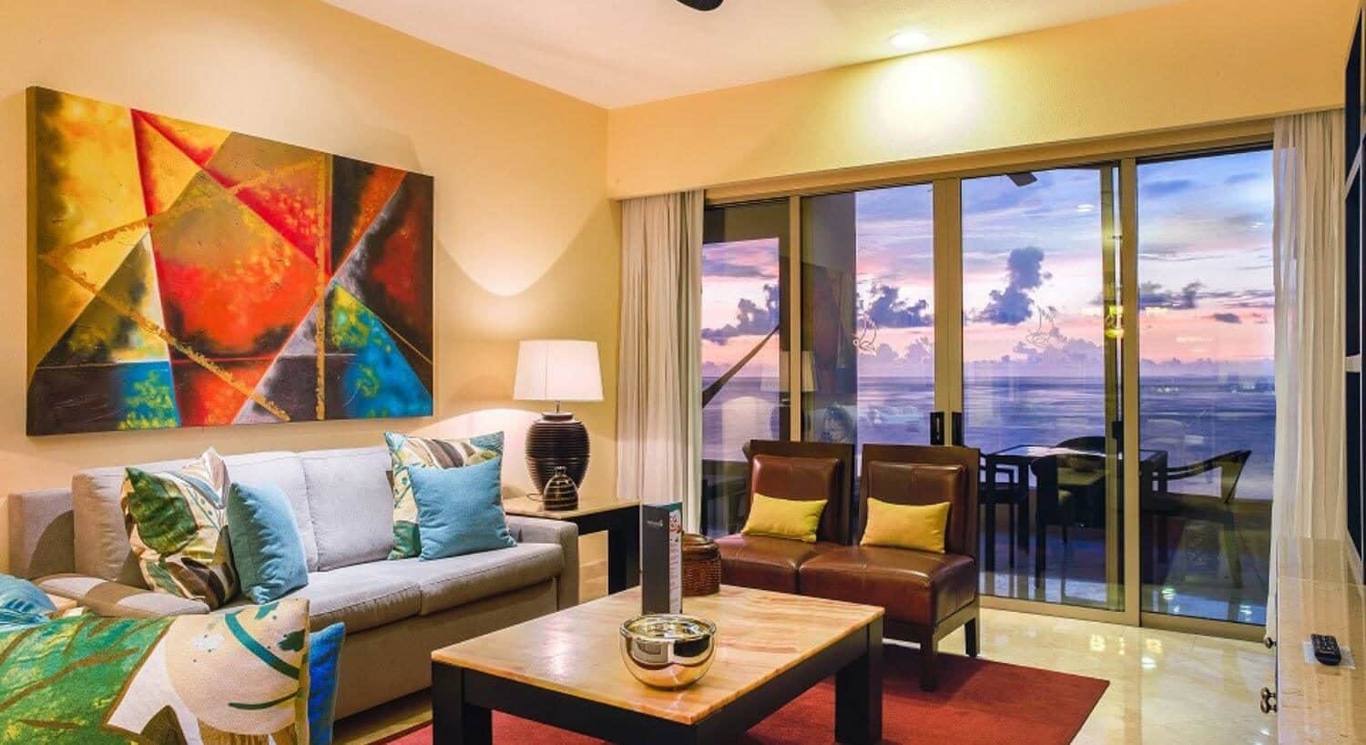 A living room with white sofas with green and blue throw pillows, two leather chairs with yellow throw pillows, marble coffee table and end table with a lamp, and sliding doors leading out to a private balcony with outdoor dining furniture and the purple and pink sunset over the ocean.