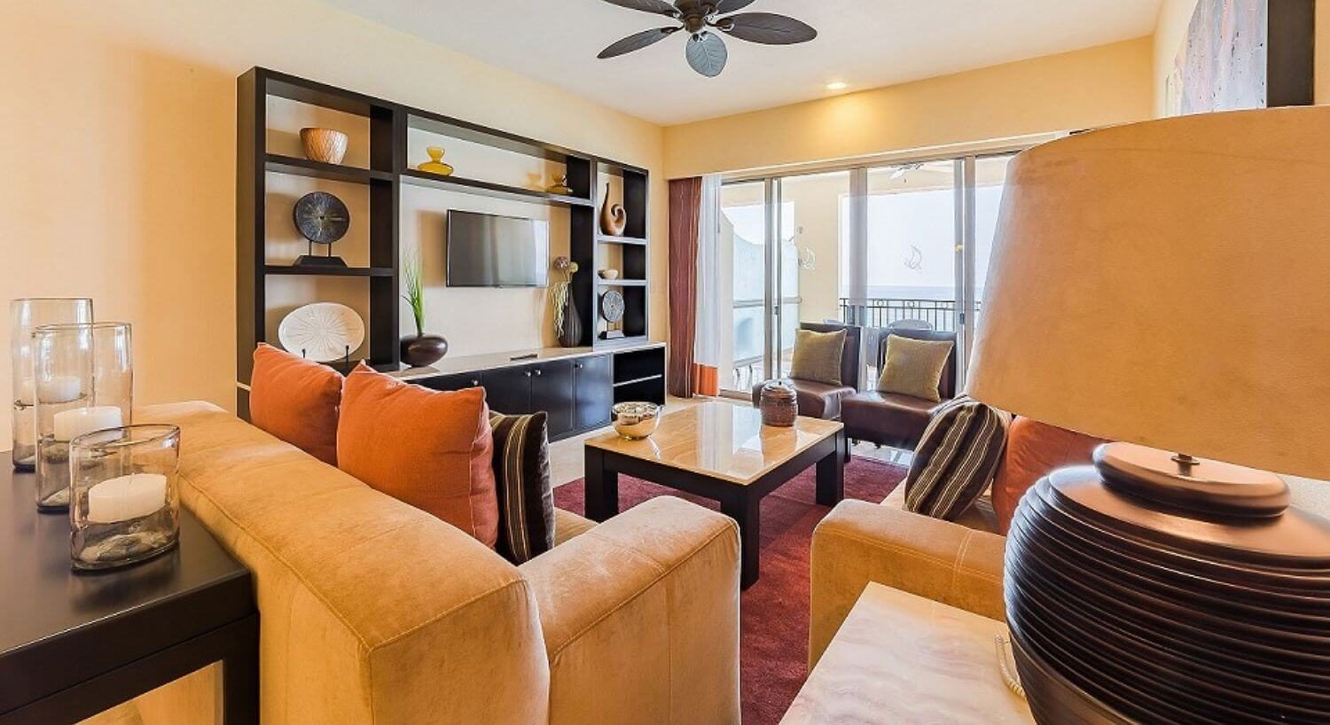 A living room with comfortable plush tan sofas and leather chairs with orange and brown accent pillows, marble coffee table and side table with lamps, an entertainment center with flat screen TV and shell and pottery accents, and sliding doors leading out to a balcony with ocean views.