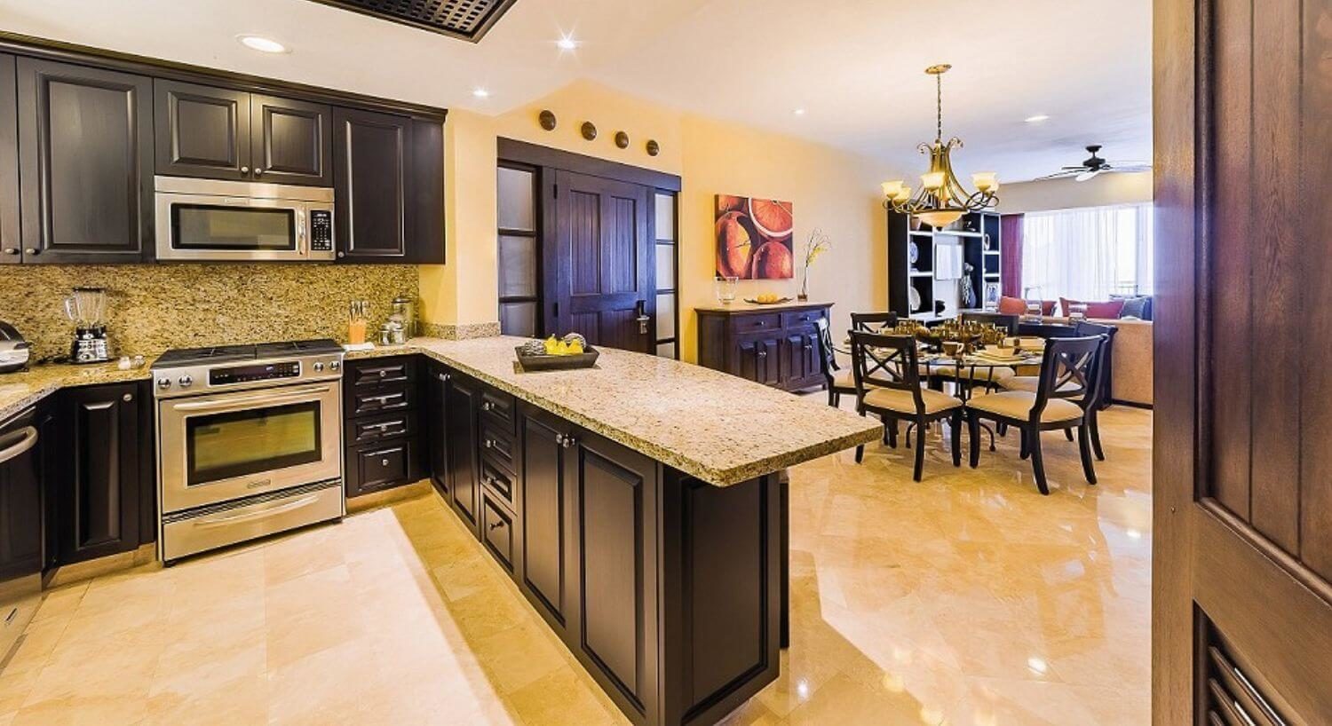 A large U shaped kitchen with stainless steel appliances. dark brown cupboards, granite countertops, a dining area with a round glass table and brown and dark brown chairs with plush tan cushions, a matching sideboared, and living room, and sliding doors leading out to a patio.