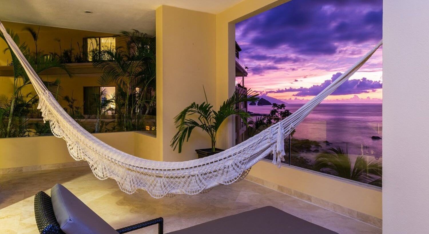 A private balcony with lounge chair and hammock, overlooking the purple sunset over the ocean and another hotel building.