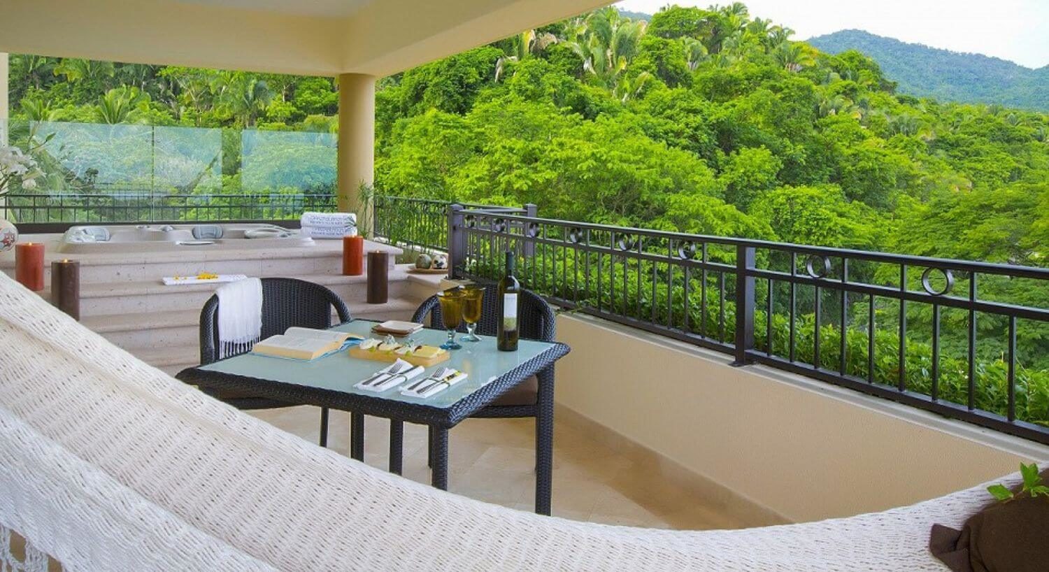 A private balcony with a hammock, dining table and chairs, hot tub, and the lush greenery of the mountain side.