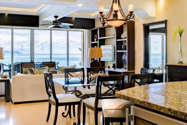 An open room with a breakfast bar, dining table and chairs, living room with sofas and chairs, and sliding glass doors that lead out to a private balcony with outdoor dining furniture, and views of the ocean.