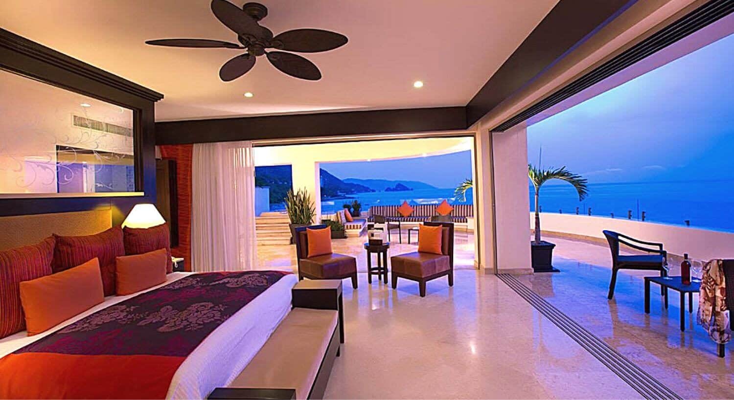 A large bedroom with a King bed, sofa, plush chairs, and open floor to ceiling windows on two sides that open to a wrap around balcony featuring outdoor living area and stunning views of the ocean and mountains.
