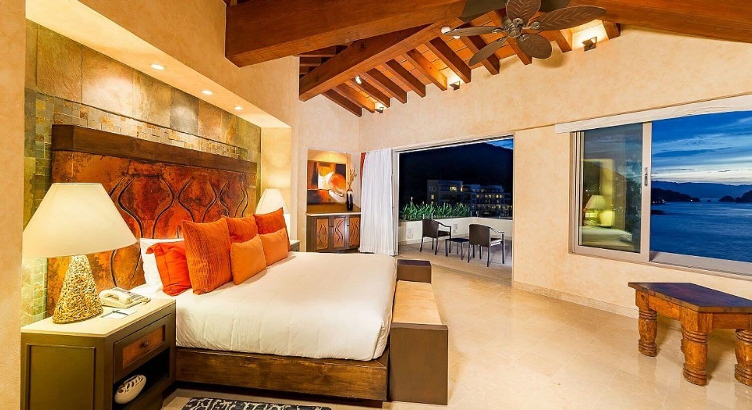 A spacious bedroom with King bed with white and orange bedding, a nightstand with lamp on either side, a bench below an open window, and sliding doors that lead out to a private balcony with patio furniture, and views of the ocean and mountains.