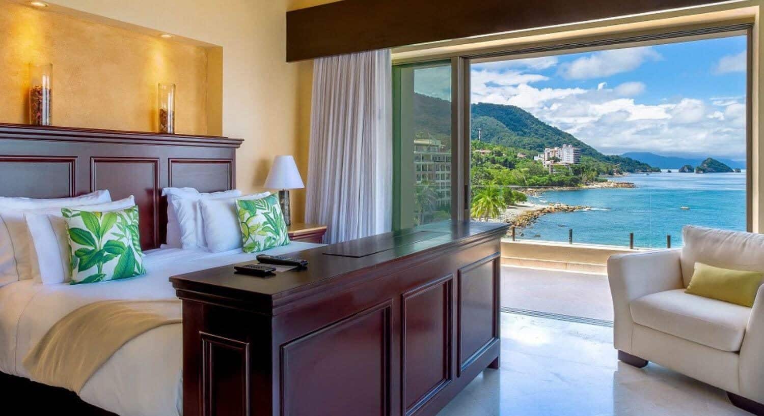 A bedroom with a King bed with wood headboard and footboard, plush armchair, and open sliding doors leading to a balcony with ocean and mountain views.