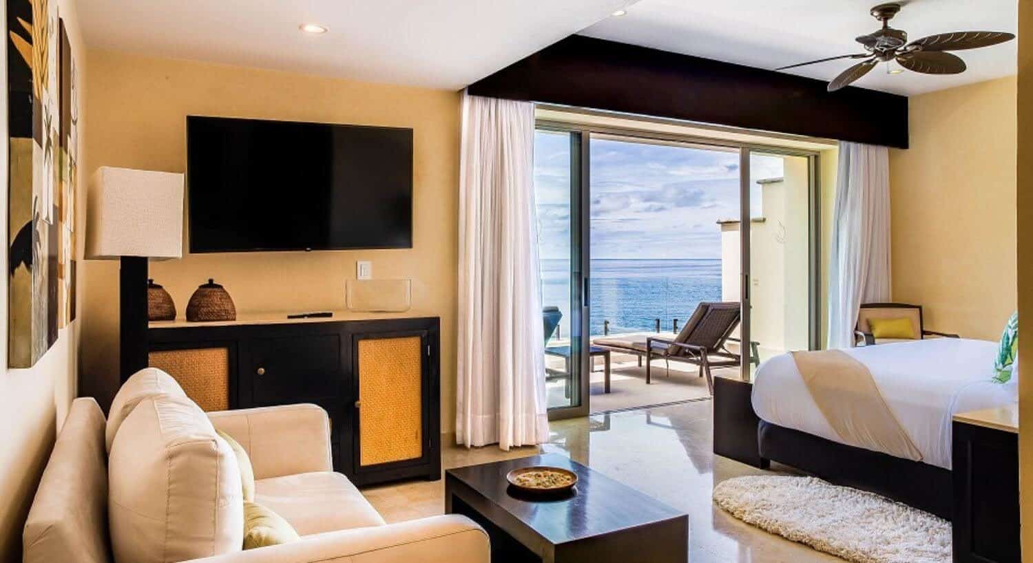 A spacious bedroom with a King bed, loveseat, flat screen TV, dresser, and sliding doors that lead to a balcony with lounge chairs, and views of the ocean.