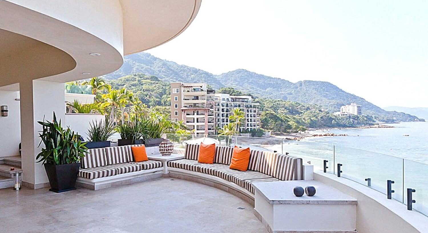 A large outdoor living area at a private penthouse with outdoor brown and white striped sofas with orange throw pillows, with views of the ocean, mountains, and other hotel buildings.