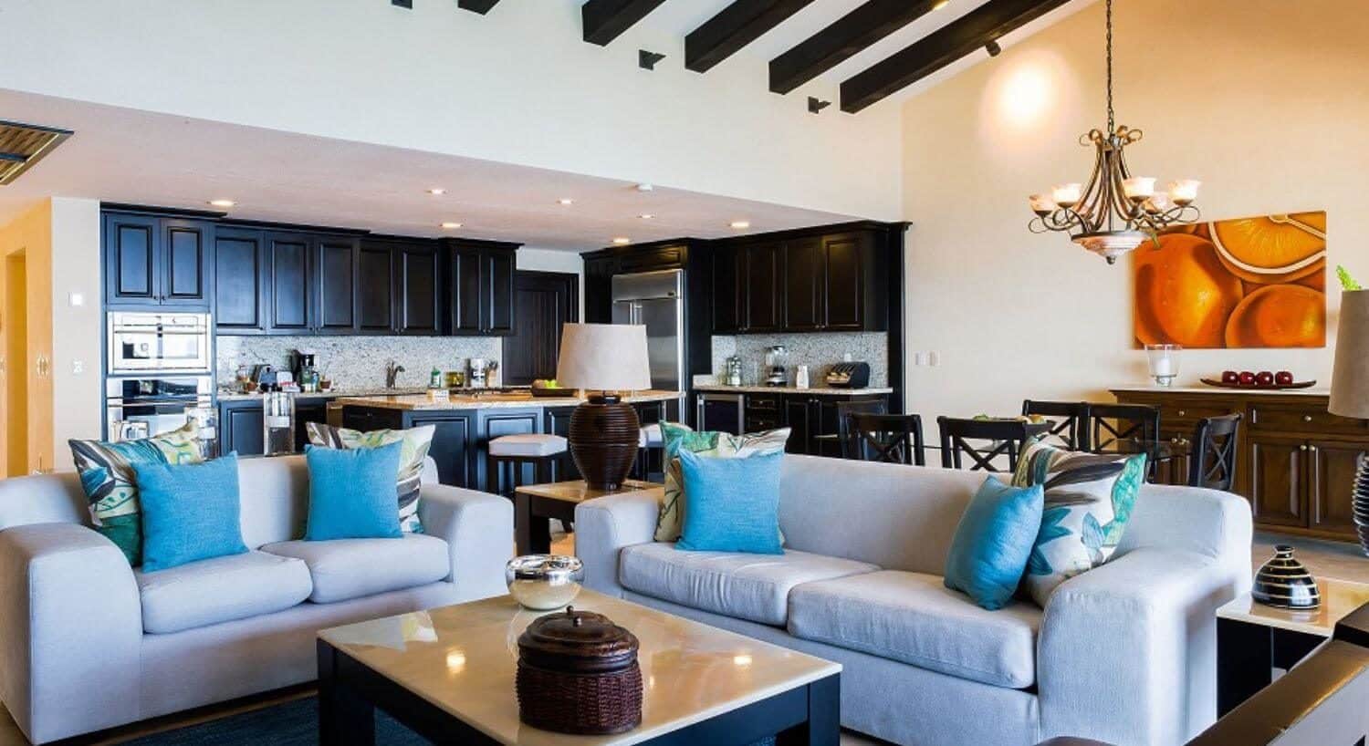 An open floor plan living area with plush white sofas with blue and green throw pillows, a marble coffee table, a dining table and chairs, and a full kitchen.