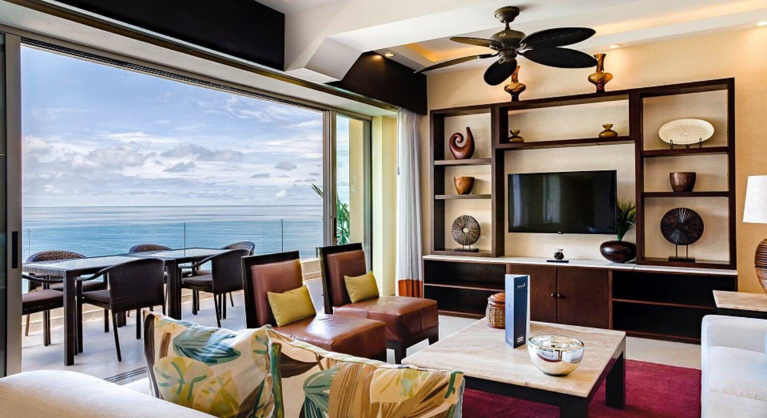 A living room with sofas and plush leather chairs, an entertainment center with flat screen TV, and floor to ceiling sliding doors that lead to a balcony with patio furniture and views of the ocean.