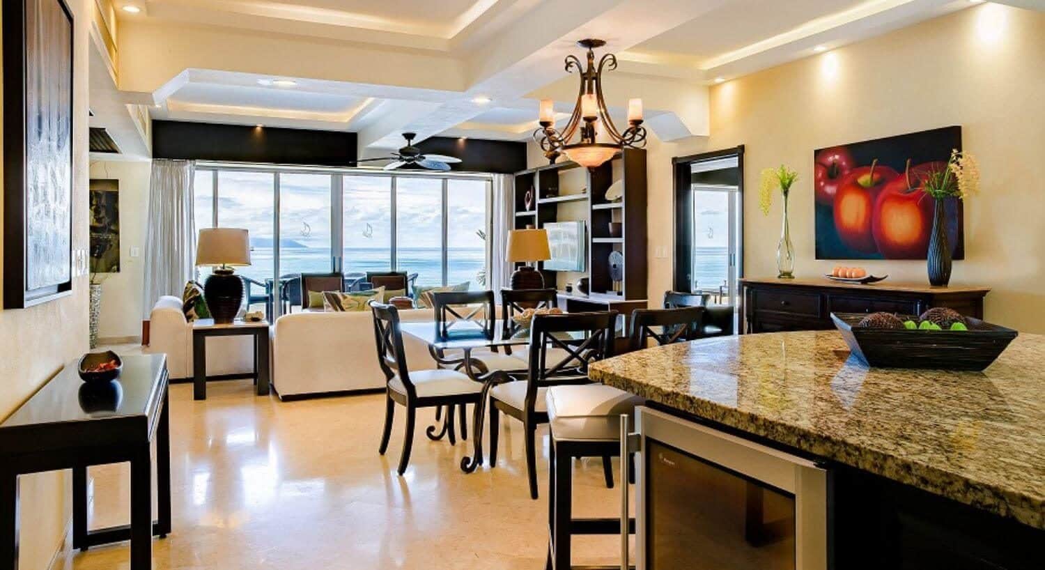 An open living and dining area including a breakfast bar with stools, dining table and chairs, and living area with plush sofas and chairs, leading out to a balcony with outdoor furniture, and views of the ocean.