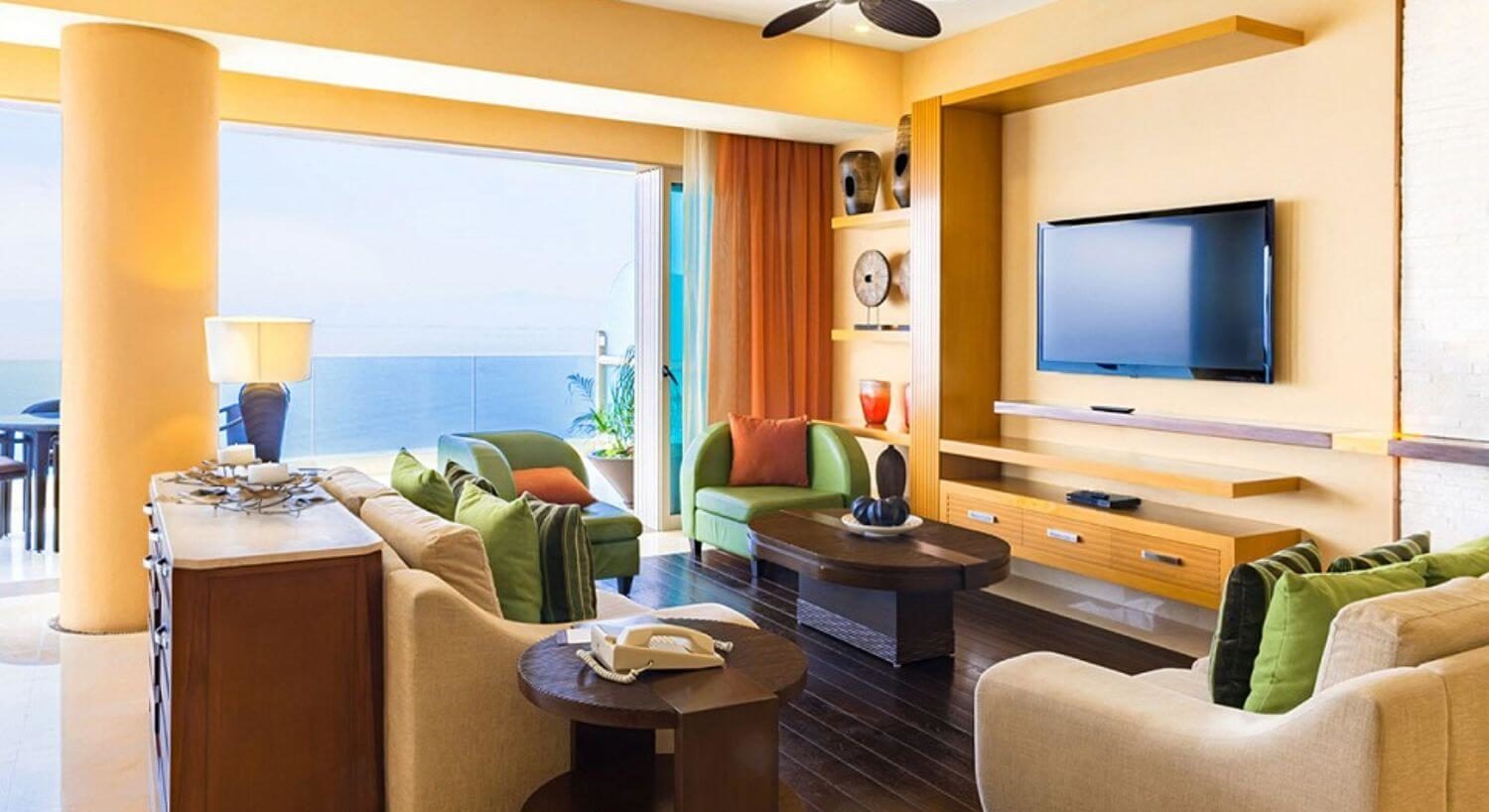 A living room with tan and green furniture with orange, brown and green throw pillows, wood coffee table and side tables, a flat screen TV and sliding doors open to a patio with ocean views.