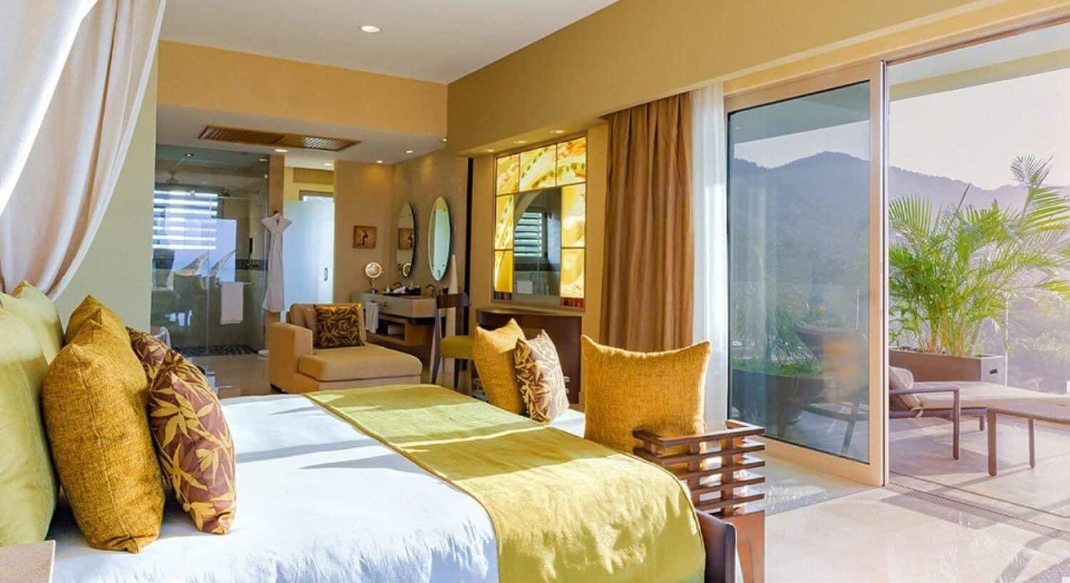 A bedroom with King bed with white, yellow and brown bedding, a plush chaise lounge chair, vanity table with lit mirror, bathroom with shower, and sliding doors leading out to a private balcony with lounge chair, green palm bushes, and views of the mountains.