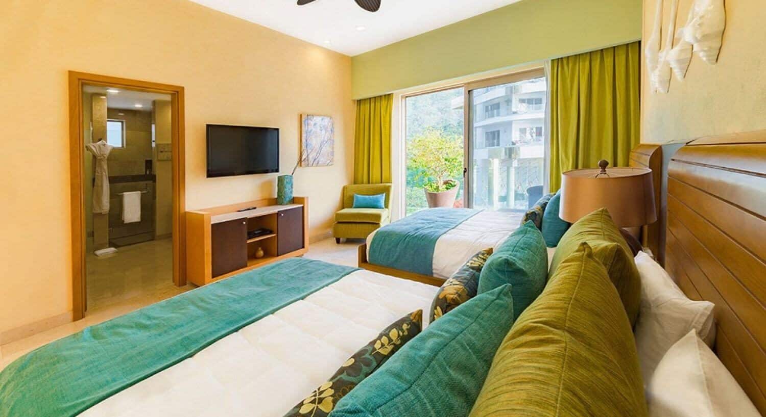A bedroom with 2 queen beds with white and green bedding, a dresser and flat screen TV, a plush green chair in the corner, sliding doors that lead out to a private patio, and a bathroom with a shower and bathrobe hanging next to the shower.