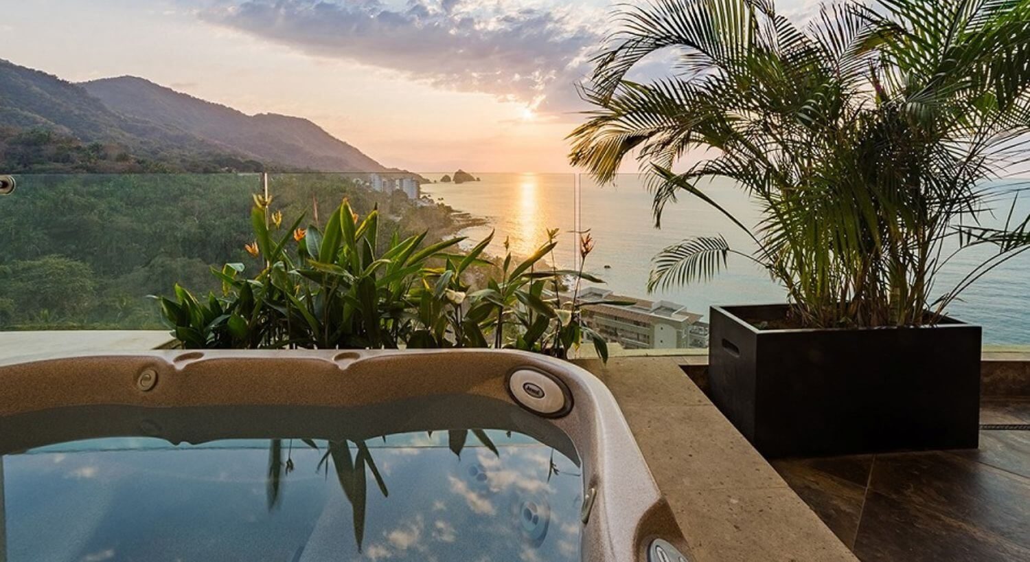 An outdoor hot tub on a balcony high above ground in the mountains with stunning sunset views of the ocean and mountains.
