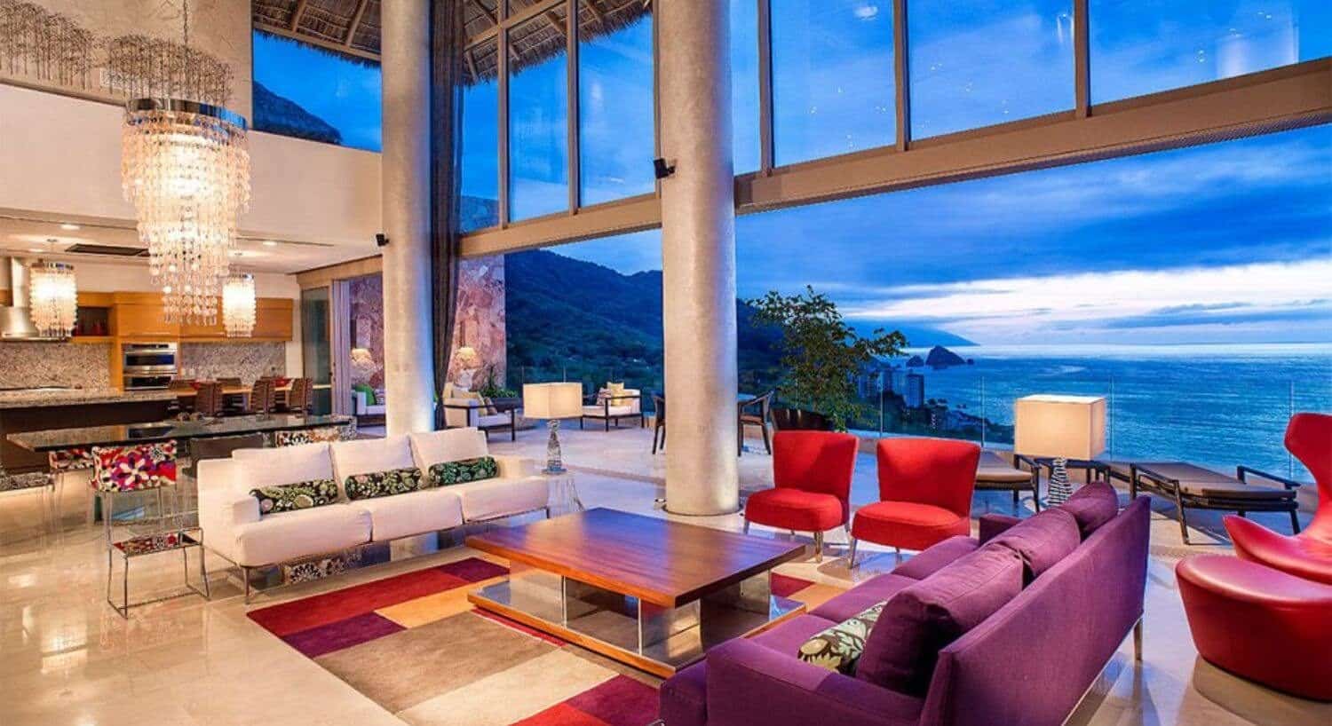 A large room with a living area of white, red, and purple furnishings, a dining table and chairs, a kitchen, and open windows all the way across the entire area opening out to an outdoor living area with stunning views of Banderas Bay.