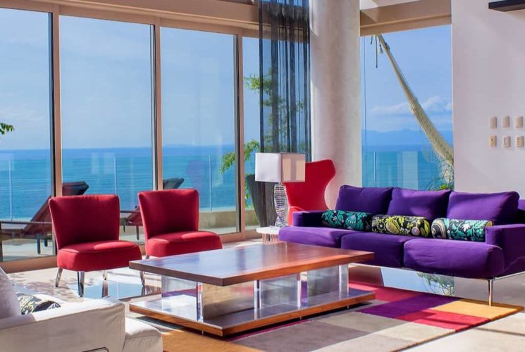 A multi story living room with plush purple, red and white furnishings and floor to ceiling windows with views of the ocean.