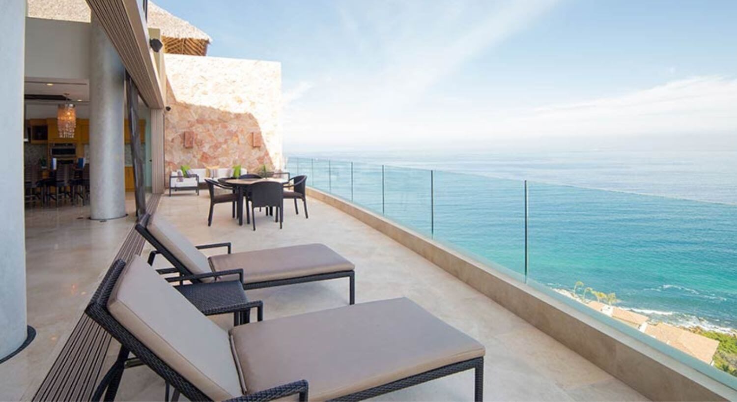 And outdoor balcony with lounge furniture that opens up to the living area of a hotel room, with views of the turquoise ocean waters of Banderas Bay.