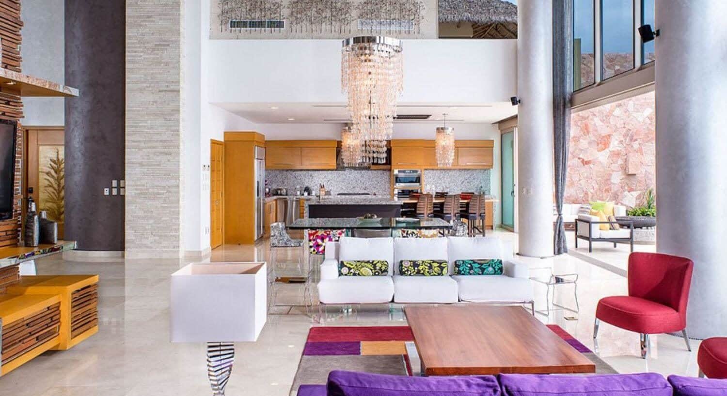 A large open room with a living area with white, purple and red furnishings, a dining area, and large kitchen, opening up to an outdoor living area.