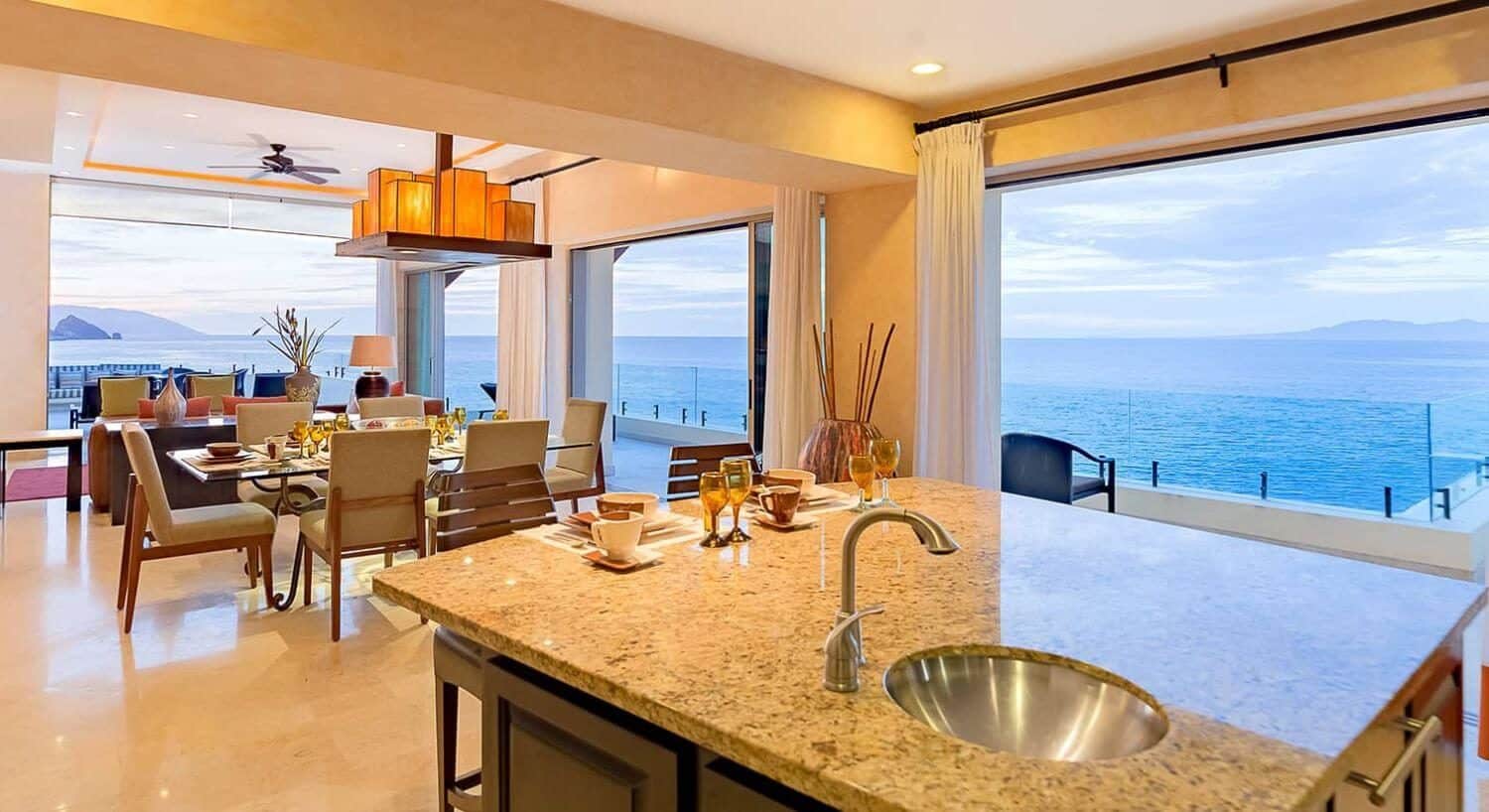 A kitchen island with sink and dining room table and sliding doors opening up to a wrap around terrace with stunning ocean views.
