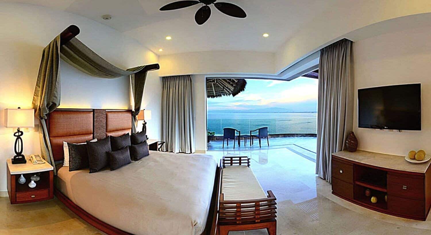 A spacious bedroom with King bed with canopy of green curtains, brown accent pillows, nightstands with lamps on either side, a cushioned bench at the foot of the bed, a dresser and flat screen TV, and sliding glass doors opened to a balcony with table and chairs facing the ocean.