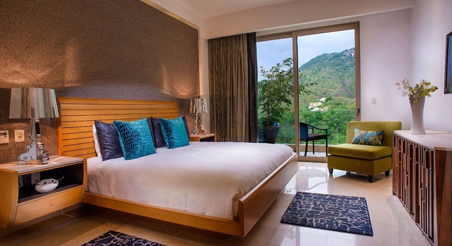 A bedroom with King bed, white beddings with turquoise and deep blue pillows, nightstands on either side with lamps, a dresser, plush armchair, and sliding doors that lead out to a balcony with views of the mountain side.