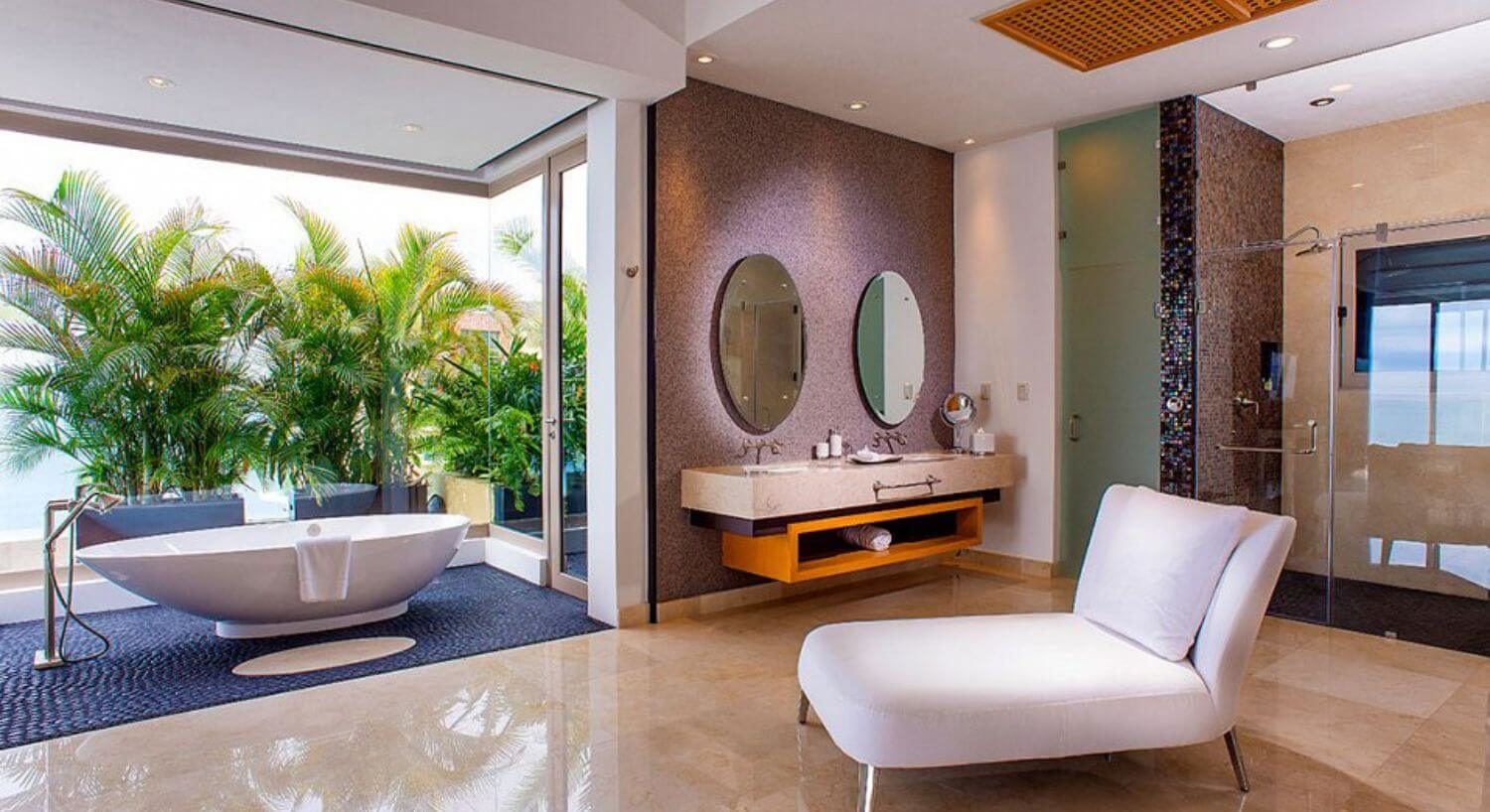 A spacious bathroom with dual sink vanity and mirrors above each sink, a plush white lounge chair, a walk in shower, a teardrop shaped bathtub set on a rock floor with windows overlooking the ocean and palm trees.