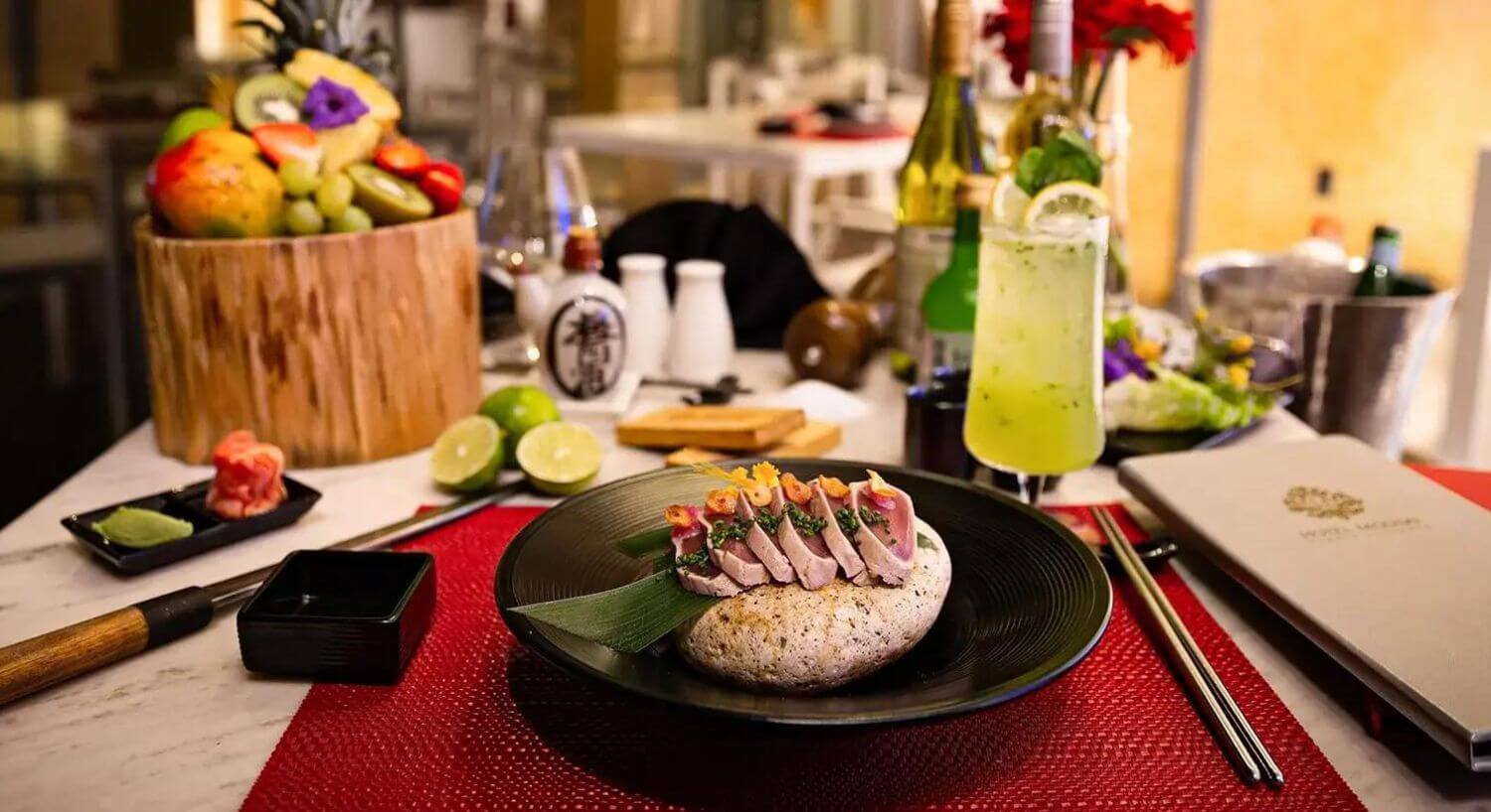 a black plate with a piece of bread and green leaf topped with seared ahi tuna, garnished with caviar and herbs, along with a green mojito cocktail, chopsticks, a menu, and a wood bowl full of fruit.