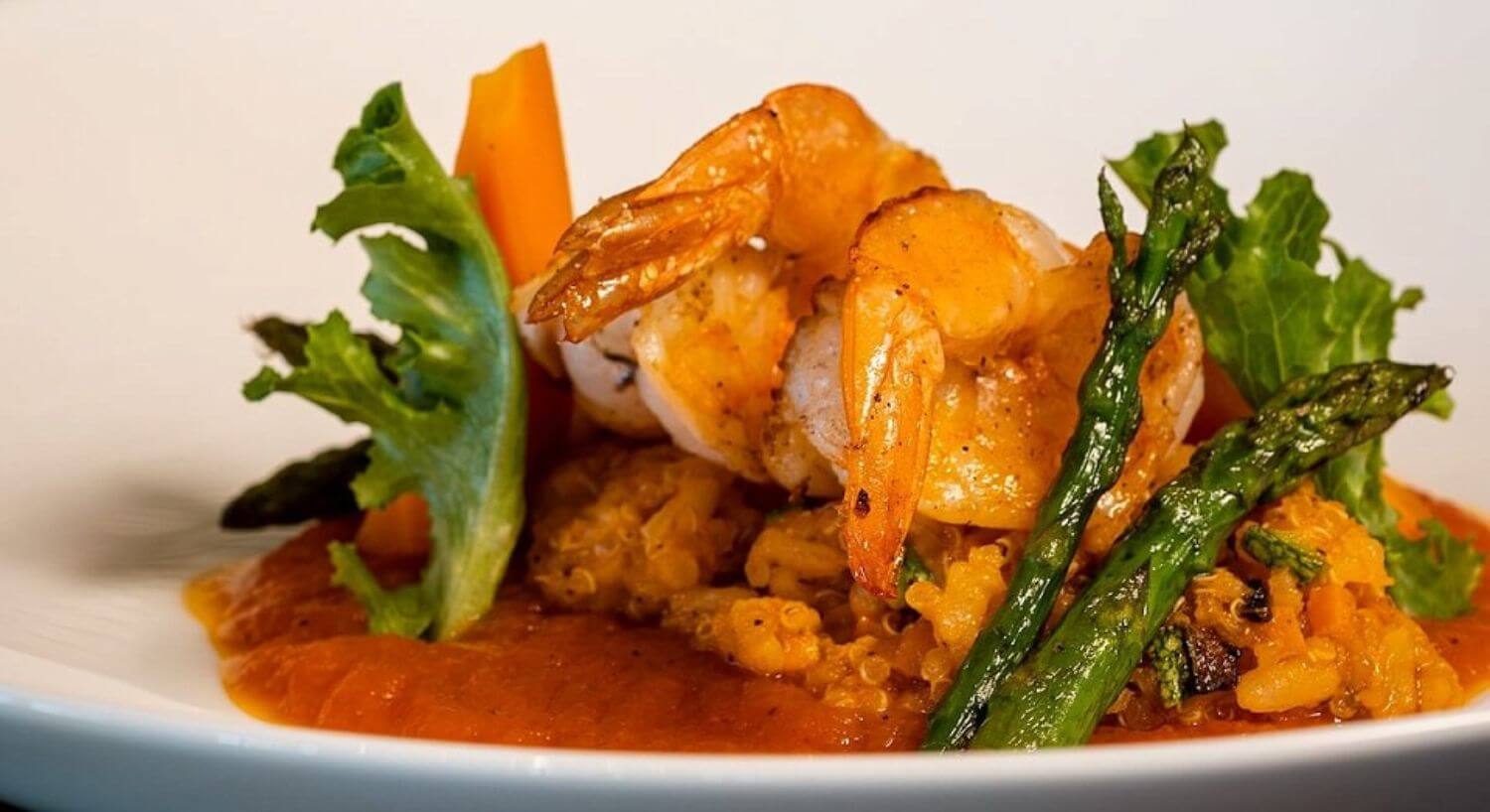 a plate with grilled shrimp, set atop rice and quinoa on a red sauce, garnished with green leaves and asparagus.