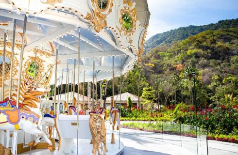 A gold and white carousel with colorful horses and chairs, set in front a lush green mountainside.