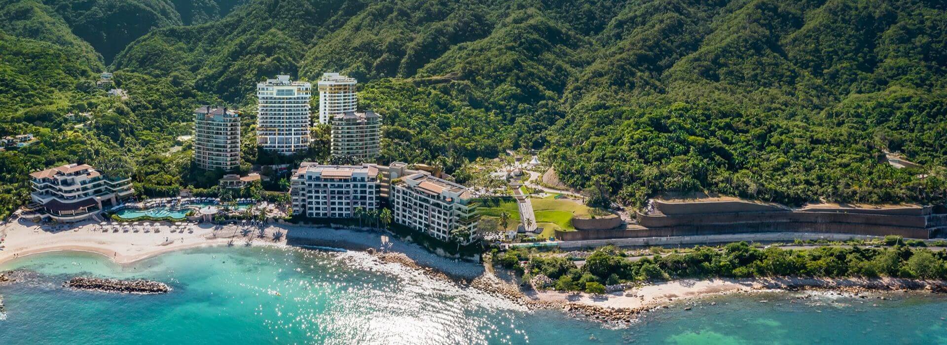 An aerial view of a lush mountainside on the ocean with several high rise hotel buildings built on the side of the mountain and on the beach front., along with a pool, a sandy beach, and the turquoise waters of Banderas Bay.