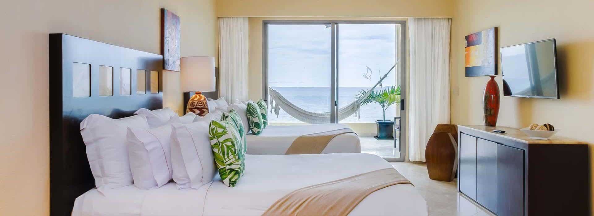 A bedroom with 2 Queen beds with white, tan and green bedding, dark wood headboard, a leather armchair, a dresser and Flat Screen TV on the opposite wall, and sliding doors leading out to a balcony with a hammock and ocean views.