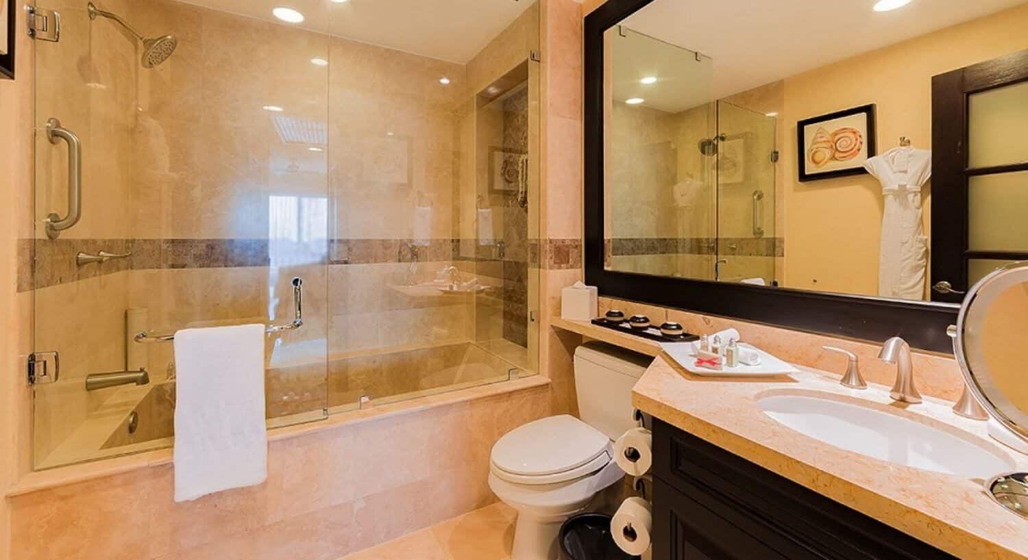 A bathroom with dark wood cupboard and marble countertop with sink, magniying mirror, bath amenities, a large mirror above, a toilet, and a large bathtub and shower with glass doors.