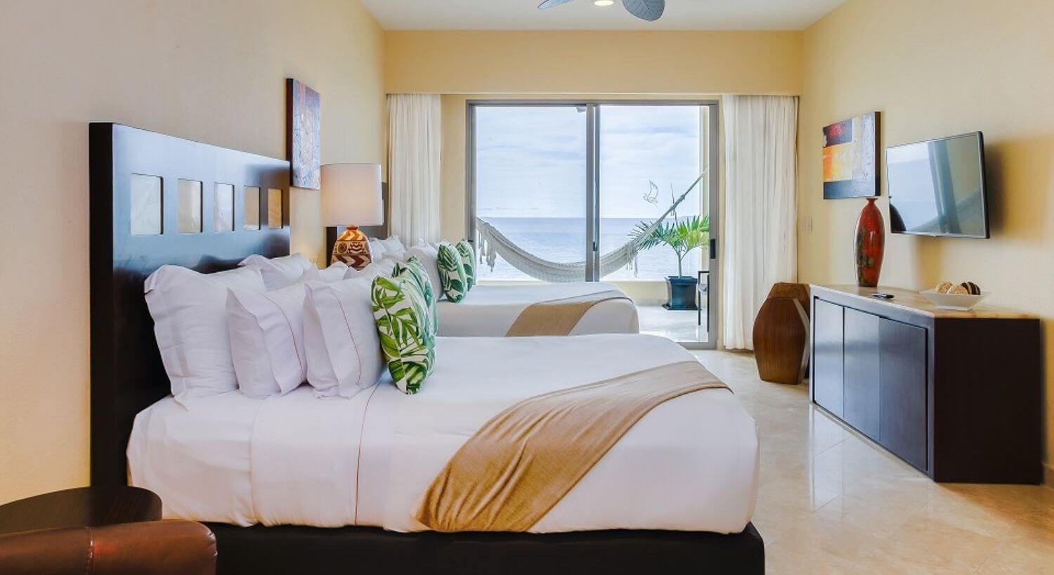 A bedroom with 2 Queen beds with white, tan and green bedding, dark wood headboard, a leather armchair, a dresser and Flat Screen TV on the opposite wall, and sliding doors leading out to a balcony with a hammock and ocean views.
