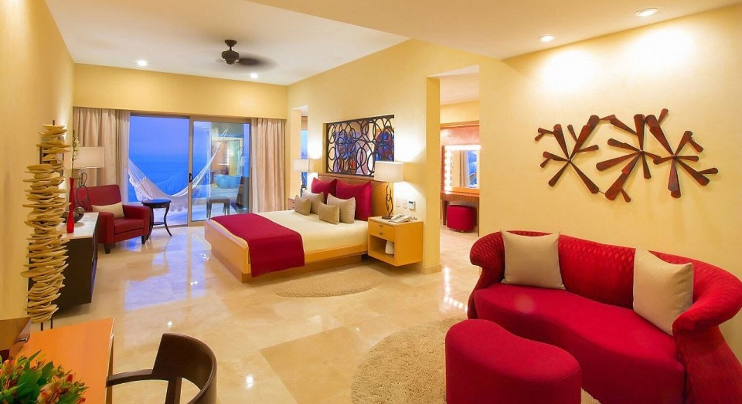A large bedroom and sitting room with a King bed with white, red and tan bedding, nightstands on either side with lamps, a red loveseat and ottoman in the sitting area with a desk and chair against one wall. Sliding glass doors open out to a private balcony with hammock and patio furniture, and beautiful ocean views.