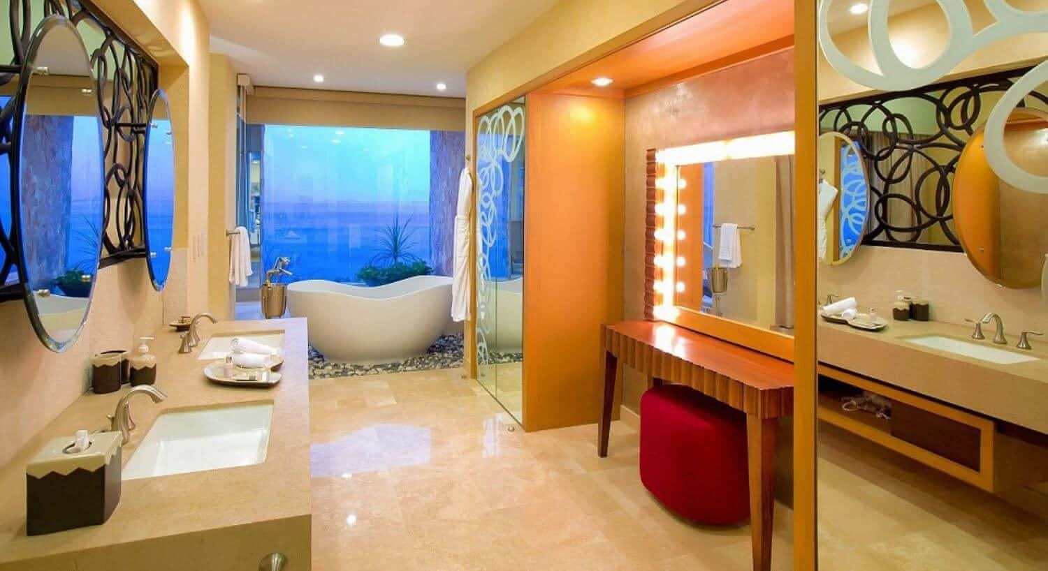 A bathroom with double sinks in a marble countertop, lit magnifying mirror, and a vanity with lit mirror and red ottoman across from the sinks. A teardrop bathtub set in rocks with floor to ceiling glass windows showcasing ocean views.