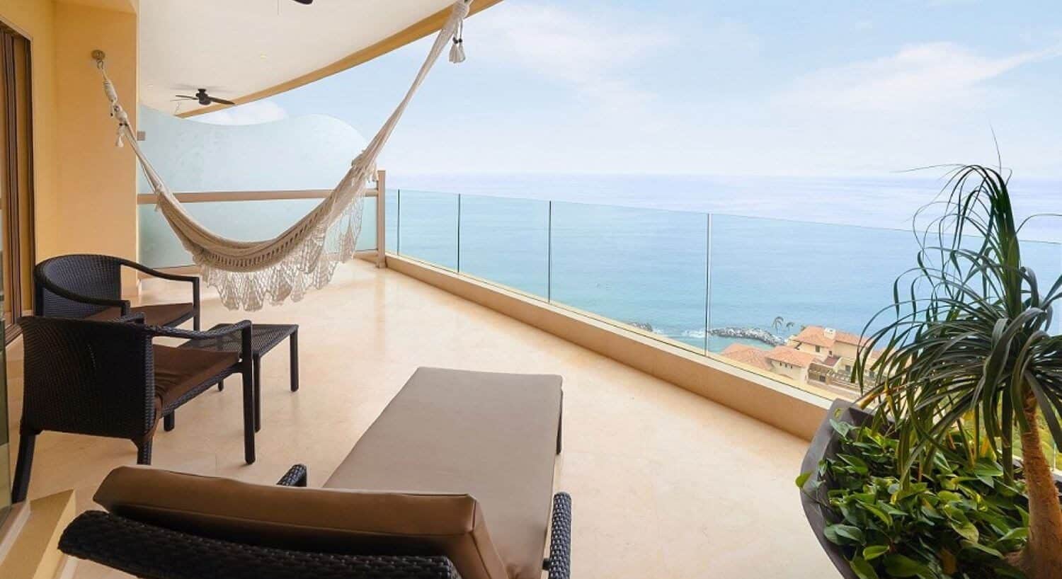 A private balcony high above the ocean with a lounge chair, patio furniture, a hammock, a planter with lush green plants, and sweeping ocean views and the tops of villas below.