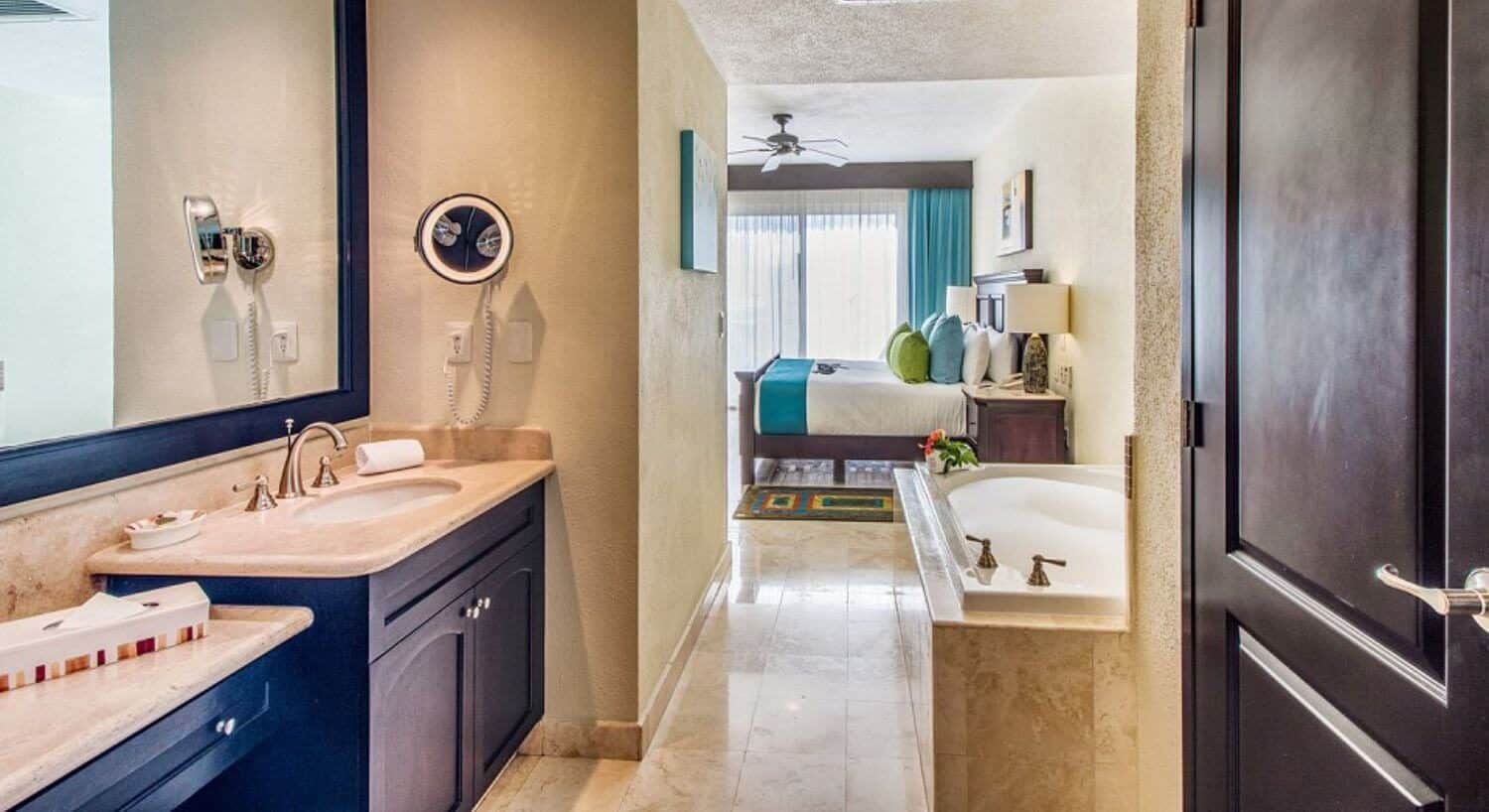 A bathroom with a sinks and vanity, dark brown cupboards, granite countertops, a deep soaking tub with bubbles; opening to a bedroom with a King bed, nightstand with lamp, and sliding doors with sheer curtains opening out to a private balcony.