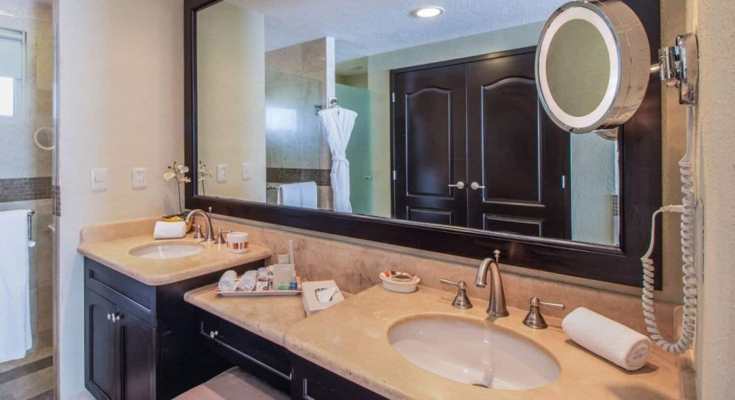 A bathroom with double sinks with a vanity with ottoman in between them, dark brown cupboards, granite countertops, and mirror the width of the sinks and vanity, and a walk in shower.