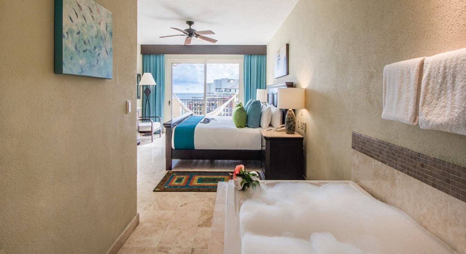 A bedroom with a King bed with white, brown and green bedding, nightstands with lamps on either side, an armchair and tall lamp in one corner, a deep tub with bubbles, and sliding doors that open to a private balcony with hammock, and views of the resort and ocean.