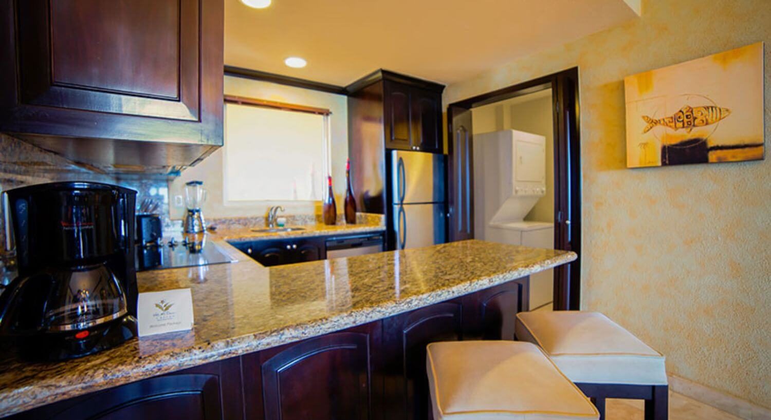 A U-shaped kitchen with dark brown cupboards, granite countertops, stainless steel appliances, coffeemaker, blender, two barstools at the breakfast bar, and an open doorway into a laundry room with stacked washer and dryer.