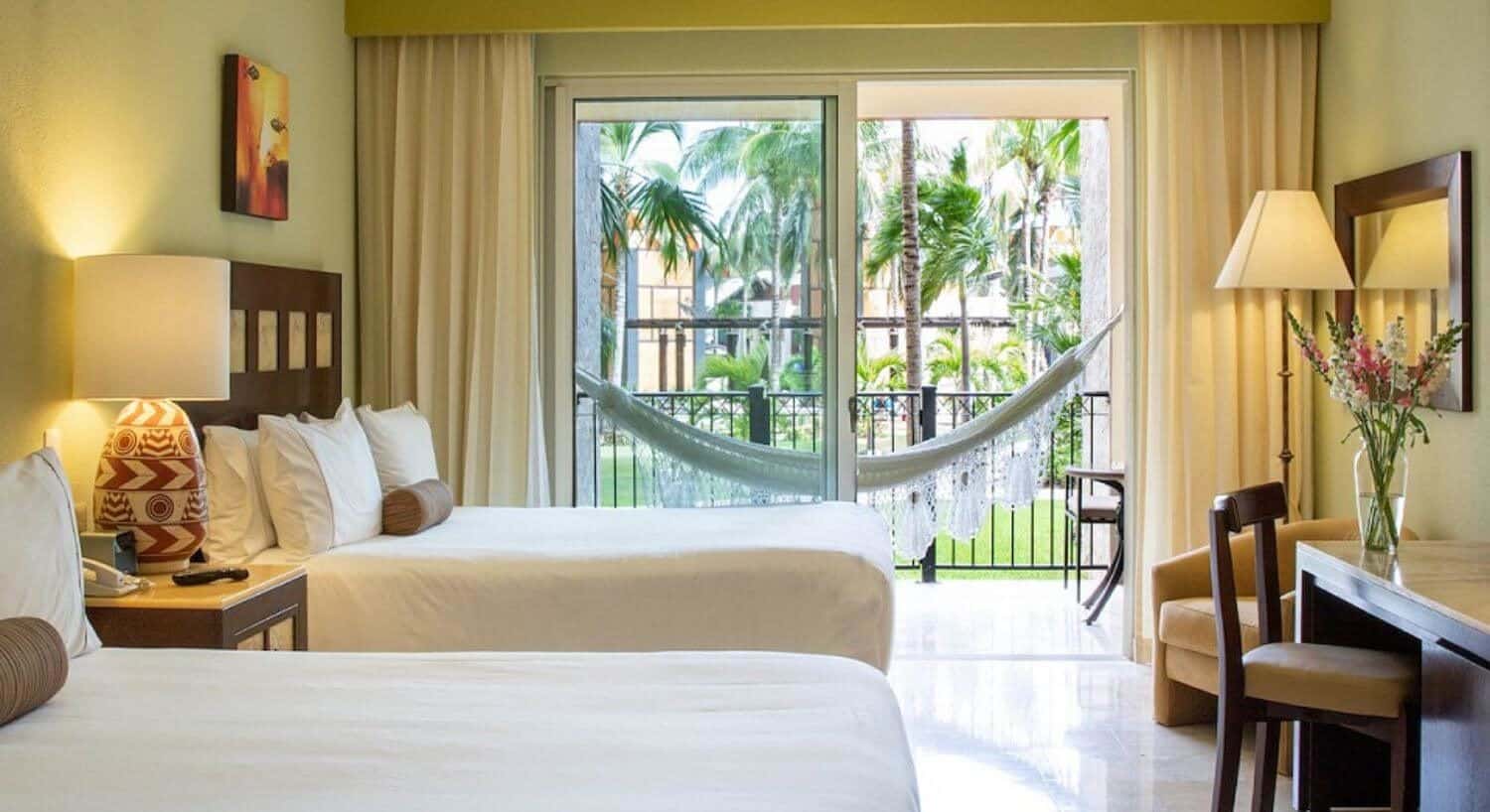 A bedroom with two queen beds with white bedding, a nightstand with lamp in between them, a writing desk and chair, a plush armchair in the corner, and sliding doors opening out to a private terrace with patio furniture and hammock, overlooking the resort grounds with green grass and palm trees.