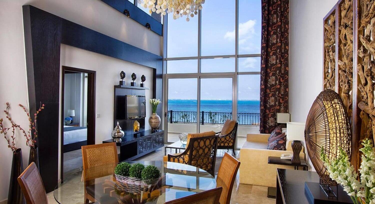An elegant living room and dining area with comfortable plush sofa and chairs, an ornate dresser with a flat screen TV on the wall, a round glass dining table and chairs, an open door to a bedroom displaying part of a bed, and multi story floor-to-ceiling windows with sliding doors opening out to a terrace with beautiful ocean views.