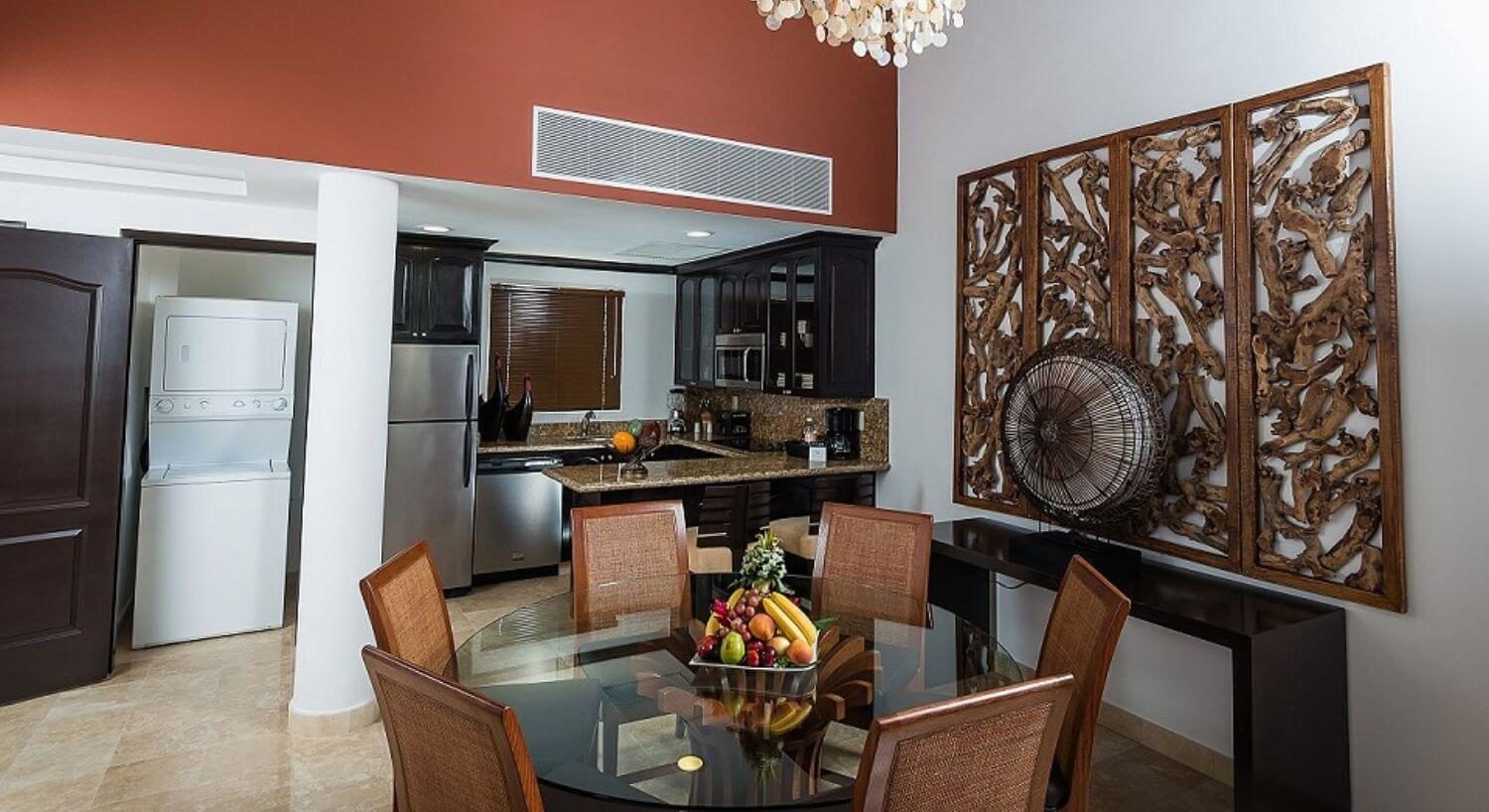 A kitchen and dining room with a U shaped kitchen with stainless steel appliances, granite countertops, dark brown cupboards, a round glass table with six chairs, and a closet with a stacked washer and dryer.