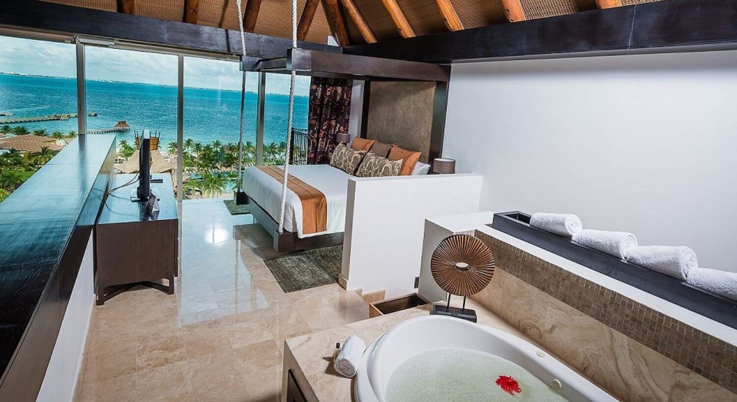 A bedroom with a seemingly hanging king bed with white, orange and brown bedding, a dresser with flat screen TV on top, a jetted tub, and floor to ceiling windows with beautiful ocean views.