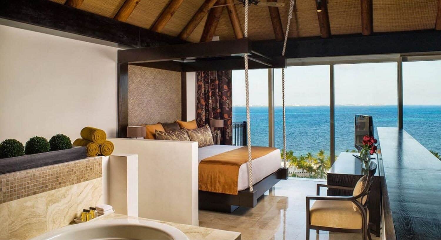A bedroom with a seemingly hanging king bed with white, orange and brown bedding, a dresser with flat screen TV on top, a plush armchair, jetted tub, and floor to ceiling windows with beautiful ocean views.