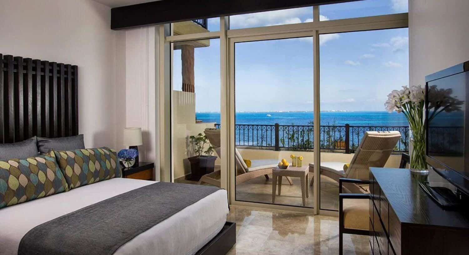 A bedroom with a king bed with white, brown and multi colored bedding, a dresser with flat screen TV on top, an armchair, and sliding doors opening up to a private balcony with lounge chairs, small table with drinks and fruit, and views of the turquoise Caribbean ocean.