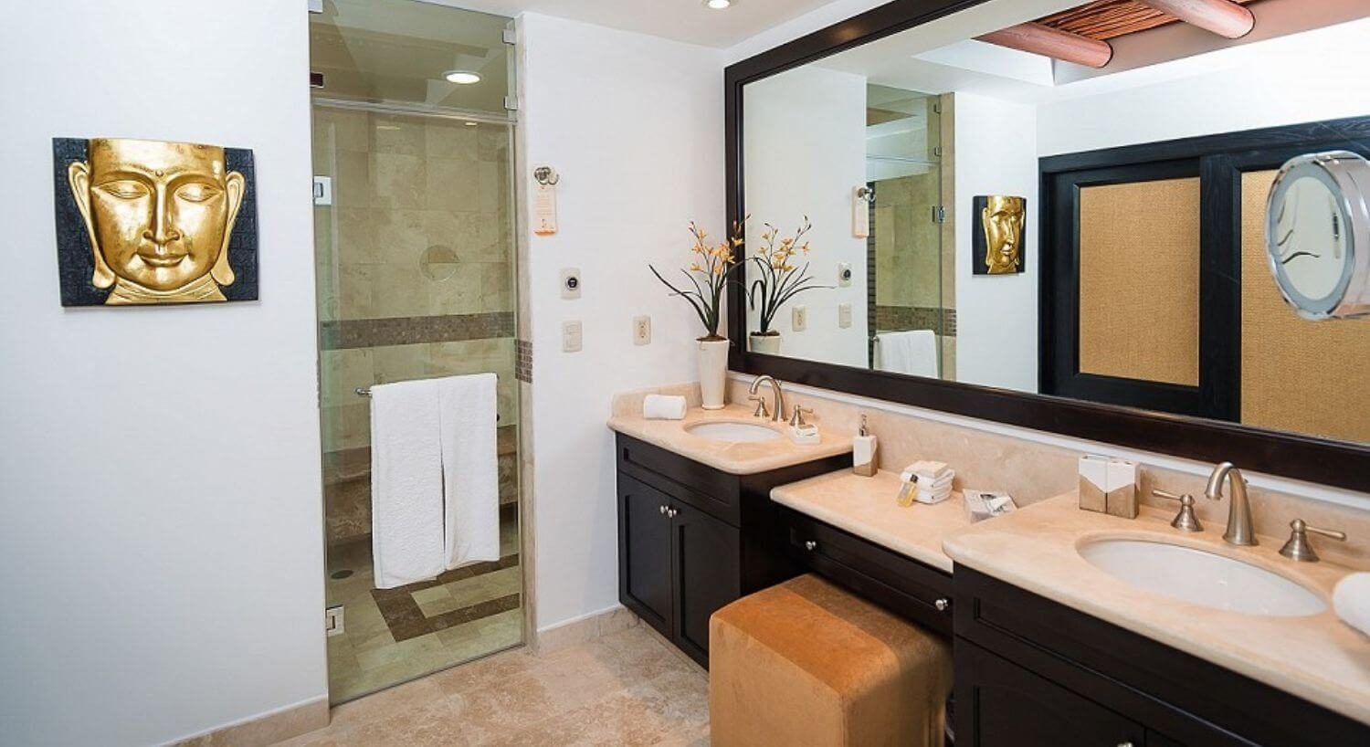 A bathroom with double vanity sinks with dark brown cupboards and granite countertops, with a vanity and ottoman in between them, and a walk in shower with clear glass door and white towels.