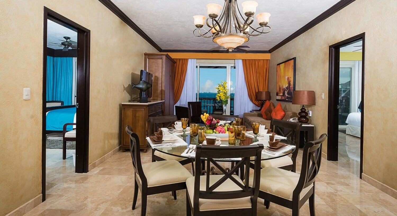 A spacious dining are with a circular glass table and several plush dining chairs, a living area with sliding doors out to a patio, and doors on either side of the dining room leading to two separate bedrooms.