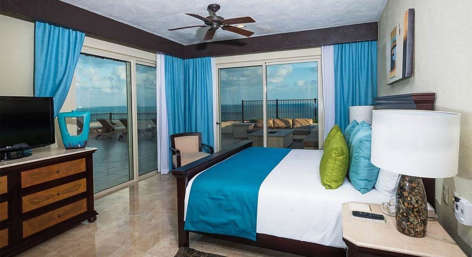 A bedroom with King bed with white, lime green and turquoise bedding, night stands and lamps on either side of the bed, a dresser and flat screen TV on the opposite wall, and sliding glass doors on two of the walls leading to an expansive wrap around terrace with patio furniture, a lounge area with fire pit, and beautiful ocean views.